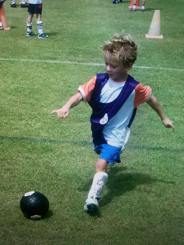 Erik as a young boy in a purple bib and blue shorts, running to kick a black ball on a grass pitch.